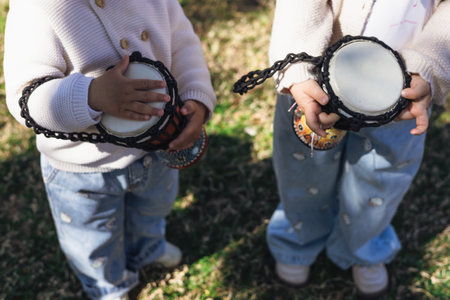 Two children in cozy sweaters play with handheld drums in a green outdoor setting. They are focused on creating rhythmic beats while smiling, enjoying a fun musical activity together.の写真素材