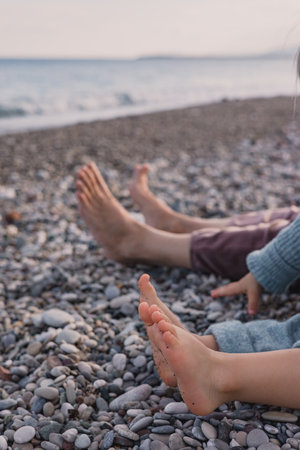Family enjoys peaceful moment by the beach, with adults and children stretching their bare feet on the smooth pebbles. The gentle waves and soft sunset create a serene atmosphere. Legs without shoesの写真素材