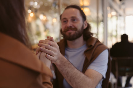 A man with a beard chats with someone in a relaxed cafe setting. He sits at a table, displaying a friendly demeanor, while patrons socialize in the background.の写真素材