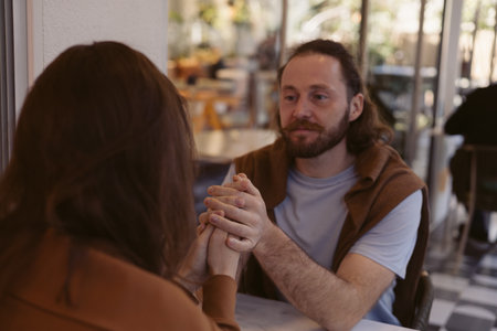couple sits at a small table in a cozy cafe, holding hands. The warm ambiance is enhanced by soft lighting while they share a tender moment, seemingly lost in conversation. Romantic date, special dayの写真素材