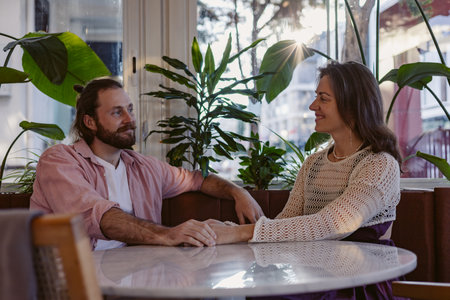 woman and man sit across each other at small table in cozy cafe. They are deeply engaged in conversation, with thoughtful expressions. Casual sporty outfit, lifestyle moments and emotions conceptの写真素材