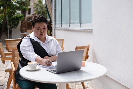 A young plus size asian woman is seated outside a cafe, focused on her laptop. Surrounded by...