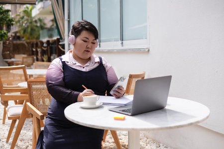 Portrait of young plus size Asian woman sits at cozy outdoor cafe, sipping coffee. She is taking notes in notebook while using her phone, laptop and listening headphones music, podcast or audiobook.の写真素材