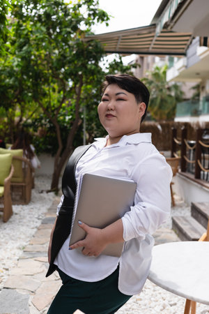 A young plus size Asian woman is engaged with her laptop outside at a cafe, smiling and concentrating. Positive female online working or studying, blogging, gadgets and educationの写真素材