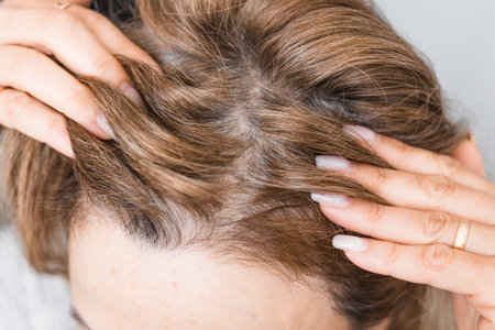 Mature middle-aged caucasian woman touching her gray, silver roots hair close-up on gray tiles background, hair loss and age-related changes, naturalness and natural aging concept.の写真素材