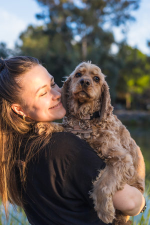 A playful moment captured as a woman joyfully holds her cocker spaniel against her chest. Their connection shines amidst lush greenery and a tranquil lake under a sunlit sky, reflecting their bond.の写真素材