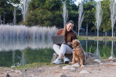 A woman sits on a rock by a serene lake, using her laptop while her cocker spaniel looks up at her....