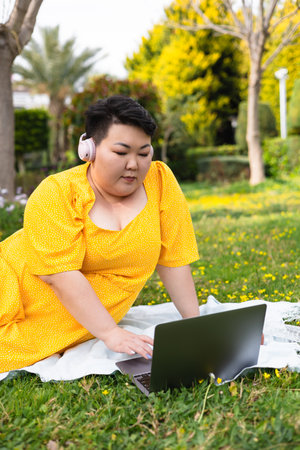 Portrait of young overweight Asian woman dressed in bright yellow dress sits comfortably on grassy area, using laptop. Sun is shining, flowers surround her. peaceful spring day filled with greenery.の写真素材