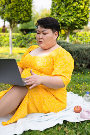 Portrait of young overweight Asian woman dressed in bright yellow dress sits comfortably on grassy area, using laptop. Sun is shining, flowers surround her. peaceful spring day filled with greenery.の写真素材
