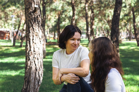 Two women mother and daughter sit on the grass in a lush park, engaged in a cheerful conversation....