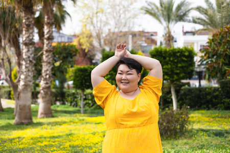 Portrait of beautiful young chubby overweight Asian brunette positive woman, female smiling, posing and walking in green park, wearing yellow dress. Sunny summer day.の写真素材