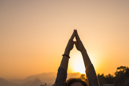 A woman stretches her arms upwards while practicing yoga at sunrise. The soft light enhances the...