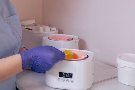 A beauty professional, wearing gloves, skillfully applies warm wax from a heater using a wooden stick, preparing for a waxing treatment. golden wax stretches from the heaterの写真素材
