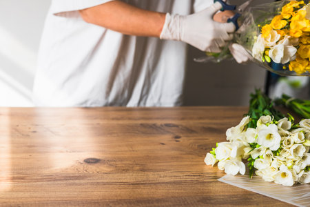 A florist carefully trims the stems of white and yellow flowers in transparent wrapping. Closeup of hands of middle-aged woman florist creating bouquet of fresh spring flowers on the table on white backgroundの写真素材