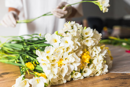 A florist is carefully arranging a bouquet of white and yellow flowers on a wooden table. Bright sunlight fills the workspace, highlighting the intricate details of the floral arrangement.の写真素材