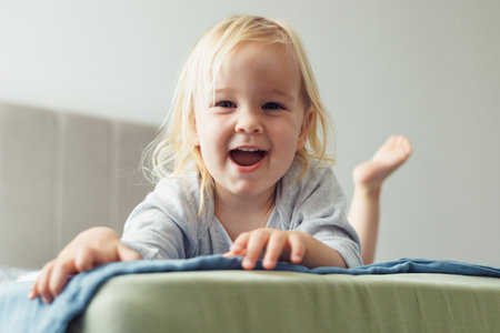 A cheerful blonde toddler is joyfully playing on a bed at home. She leans forward with a big smile, showing her playful spirit in a cozy and comfortable environment during the day.の写真素材