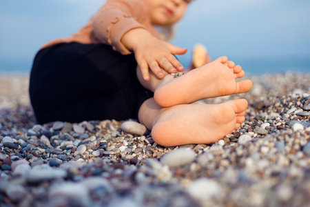 Two years girl toddler sits on a pebbly beach, playing with a yellow bucket and blue shovel. Sunlight shines on the childs bare feet and the colorful toys while waves gently lap in the background.の写真素材