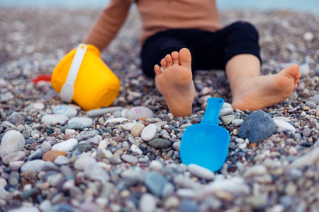 Two years girl toddler sits on a pebbly beach, playing with a yellow bucket and blue shovel. Sunlight shines on the childs bare feet and the colorful toys while waves gently lap in the background.の写真素材