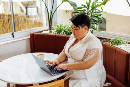Overweight Asian woman sitting comfortably at a cafe table, using her laptop while enjoying the indoor greenery.の写真素材