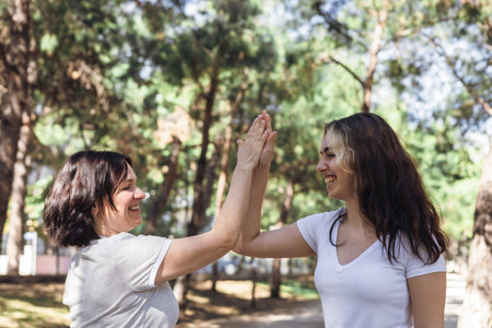 Two women mother and daughter laugh and share joyful high five in serene park. Sunlight filters...