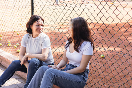 Two women mother and daughter sit on the ground beside a tennis court, engaging in a friendly conversation after training, game. They enjoy warm weather while surrounded by scattered tennis balls.の写真素材