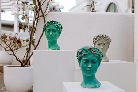 An outdoor arrangement showcases artistic busts in varying shades of green and white on white pedestals. Surrounding greenery enhances the tranquil atmosphere of this modern display.の写真素材
