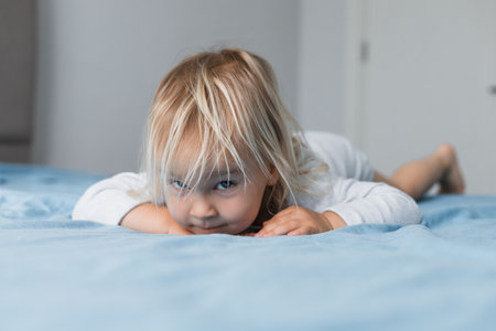 A blonde toddler girl plays on a soft bed at home, looking curiously at the camera. Her playful expression captures the joyful spirit of childhood in a cozy setting.の写真素材