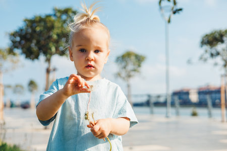 A young girl stands in a park, carefully holding dandelion seeds in her hand, enjoying a sunny day....