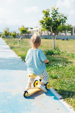 A young child enjoys riding a yellow scooter along a well-maintained path in a park. The area features lush grass and trees under a clear sky, creating a cheerful outdoor atmosphere.の写真素材