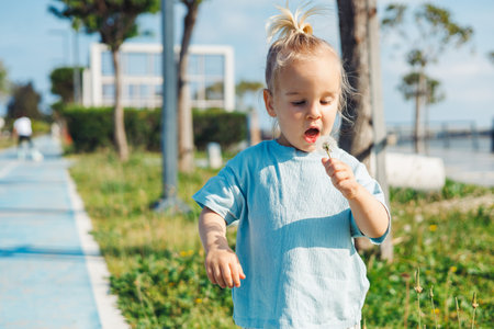 Funy young child toddler girl with open mouth stands on path in vibrant park, joyfully blowing on a dandelion flower. Hot summer day outdoor, spending time with familyの写真素材