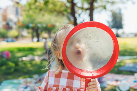A young child in a checkered orange and white dress holds a red strainer, playfully shouting into it. The setting is a lively picnic scene in a park, with green grass and blankets spread out nearby.の写真素材