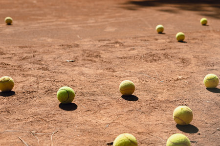 A collection of tennis balls rests on a dusty clay court under bright sunlight. The scene captures the essence of a busy practice session, with shadows indicating the time of day.の写真素材