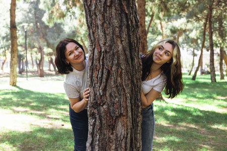 Two women mother and daughter stand playfully behind tree in a vibrant park, smiling at camera. The sunlight filters through the leaves, creating a cheerful atmosphere during their outdoor adventure.の写真素材