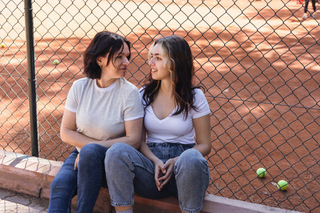 Two young women sit together against a fence at a tennis court, enjoying each others company while basking in the warm afternoon sun. Green tennis balls are scattered nearby.の写真素材