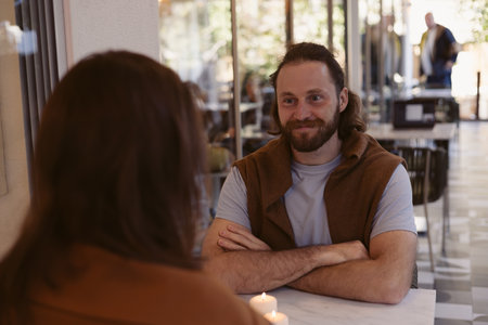 A man with a beard chats with someone in a relaxed cafe setting. He sits at a table with a candle, displaying a friendly demeanor, while patrons socialize in the background.の写真素材