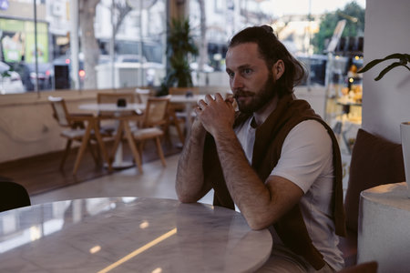 Young man sits in a cafe surrounded by lush indoor plants. The sunlight filters through the windows, creating a warm atmosphere while he enjoys his moment of reflection.の写真素材