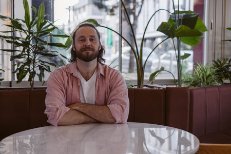 Young man sits in a cafe surrounded by lush indoor plants. The sunlight filters through the windows, creating a warm atmosphere while he enjoys his moment of reflection.の写真素材