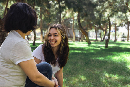 Two friends or family mature mother and teenage daughter enjoying a sunny day in a lush green park with trees in the background.の写真素材