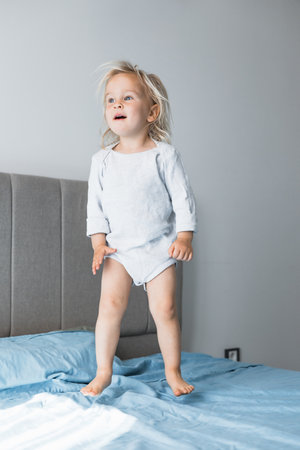 A blonde toddler girl enjoying playtime at home, energetically jumping on the bed. Soft afternoon light creates a warm atmosphere in her cozy bedroom, emphasizing her joyful activity.の写真素材