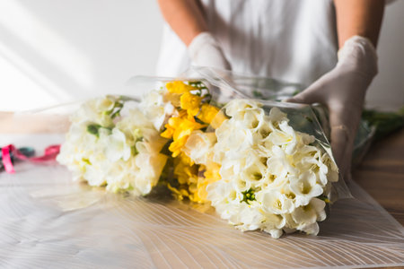 A florist, wearing gloves, is carefully arranging white and yellow flowers in wrapping paper. Scissors and ribbons are visible in the background, highlighting the vibrant workspace and creativity.の写真素材