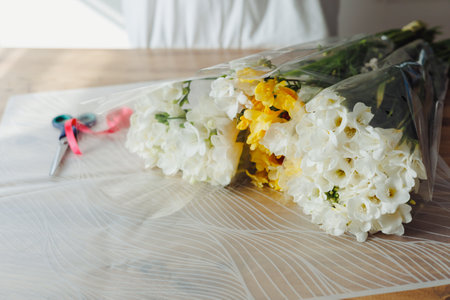 A florist with white gloves arranges vibrant white and yellow flowers, carefully wrapping them in clear film. Scissors and ribbons await on the wooden table, adding charm to the setting.の写真素材