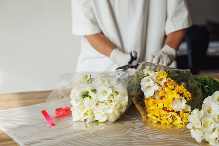 A florist, wearing gloves, is carefully arranging white and yellow flowers in wrapping paper. Scissors and ribbons are visible in the background, highlighting the vibrant workspace and creativity.の写真素材