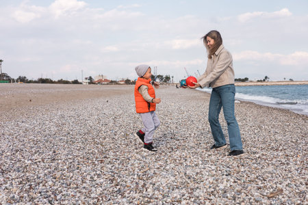 A mother and her son play together on a pebble-strewn beach during winter. They are laughing and having fun while enjoying the tranquil seaside atmosphere.の写真素材