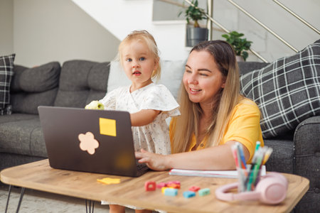 Young Caucasian woman and blond Caucasian girl using laptop at home table with school supplies and apple, online learning concept.の写真素材