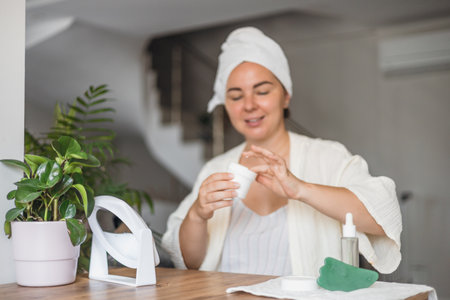 Smiling woman with a towel on her head applies cream while relaxing with a drink indoors.の写真素材