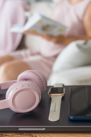 A cozy living room scene with a young woman reading a book. On a wooden table, there are pink headphones, a smartwatch, and a smartphone.の写真素材