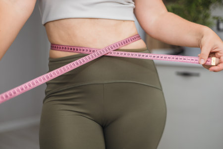 A young Caucasian woman measures her waist with a pink measuring tape. She wears a beige top and green leggings. The background is softly blurred.の写真素材