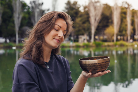 A young Caucasian woman with long brown hair holds a wooden bowl near a serene lake. Trees and greenery are visible in the background.の写真素材