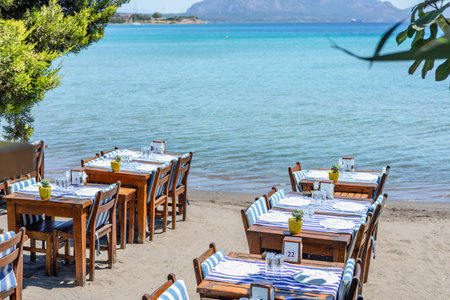 Tables lined up on the beach with turquoise water and boats in the background on a sunny day.の写真素材