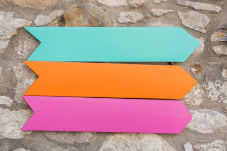Vibrant wooden arrows in pink, orange, and teal are displayed against a textured stone wall outdoors...